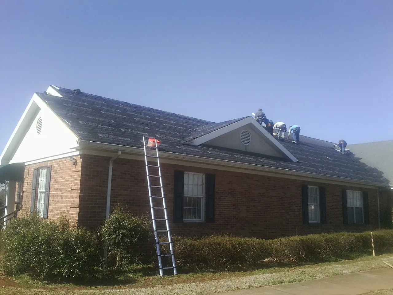 Crew tearing shingles off a brick ranch roof with a ladder set — Metal Roof Repair work by our crew in Madrid